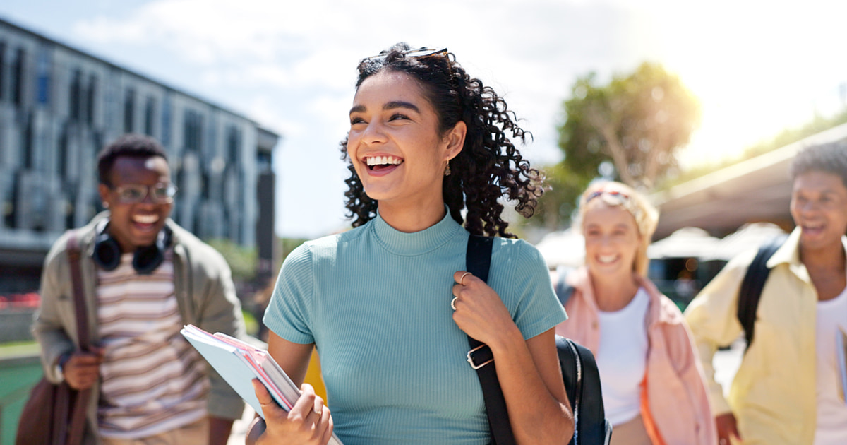 Young person walking outdoors on a sunny day, carrying notebooks and a backpack, with other students walking behind across a campus or urban walkway.