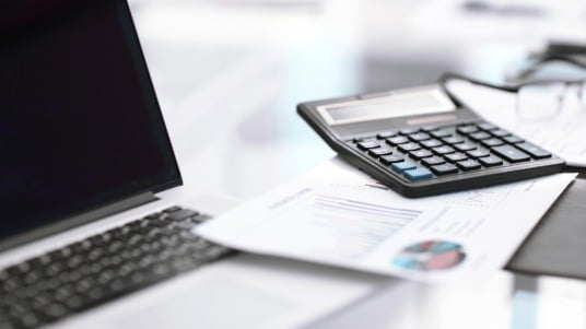 A desk surface with a laptop, a calculator and documents spread out. 