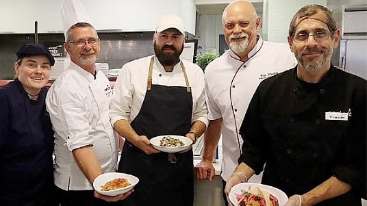 Chef Efrén Hernández with SUNY chefs presenting plated dishes in a campus kitchen. 