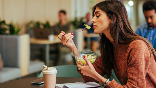 Person eating a salad at a table with a coffee cup and notebook in a casual indoor setting.