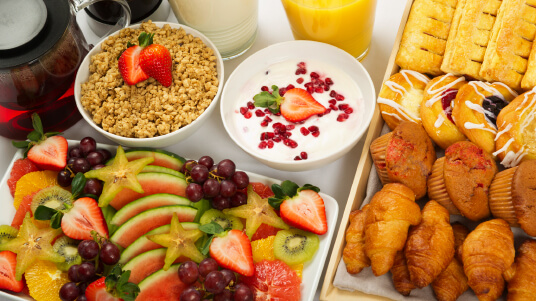 Assorted breakfast spread with fresh fruit, yogurt, granola, pastries, croissants, and drinks including juice and coffee.