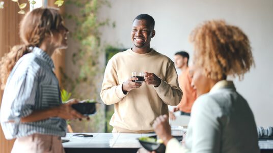 Three people standing around a counter holding bowls and cups, engaged in conversation in a bright indoor setting.