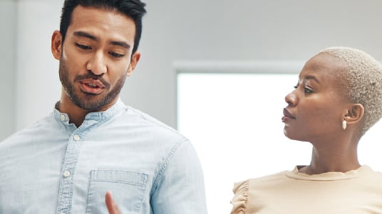 Two people in conversation indoors, one gesturing while the other looks on attentively in a bright room