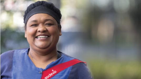 Food service worker in uniform with Sodexo sash smiling outdoors with blurred background