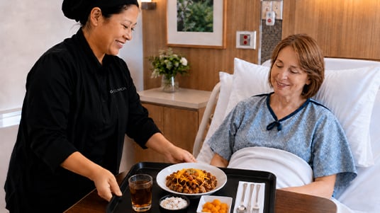 Patient receiving a meal tray from food service staff while resting in a hospital bed.