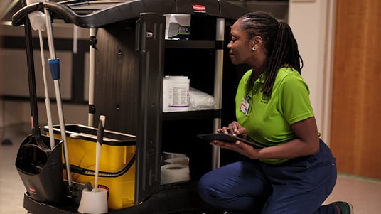 Environmental services worker inspecting and restocking supplies on a cleaning cart in a healthcare facility, highlighting sanitation operations, infection prevention, and facility support services.