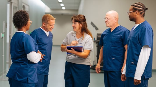 Healthcare team members gathered in a hospital corridor reviewing information on a tablet, highlighting clinical collaboration, care coordination, workforce engagement, and patient support services.