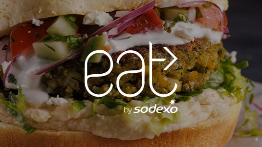 Close-up of a sandwich with vegetables and sauce, overlaid with “eat by Sodexo” logo.