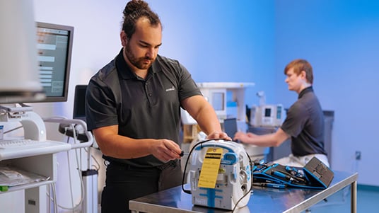 Workers testing medical equipment on a lab table, with monitors and devices in a clinical workspace.