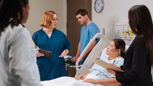 Healthcare staff gathered around a patient in a hospital bed, discussing care with charts and medical equipment visible.