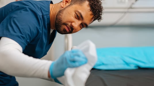 Healthcare worker in scrubs wearing gloves wipes a hospital bed with a cleaning cloth.
