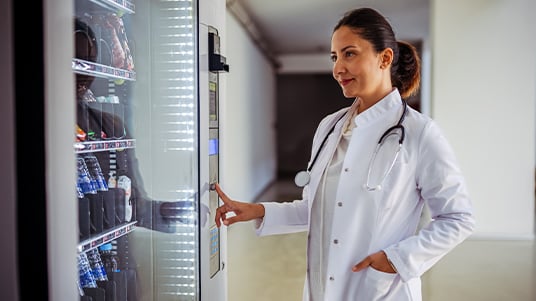Clinician in a lab coat uses a vending machine in a hospital hallway, with snacks visible behind glass.