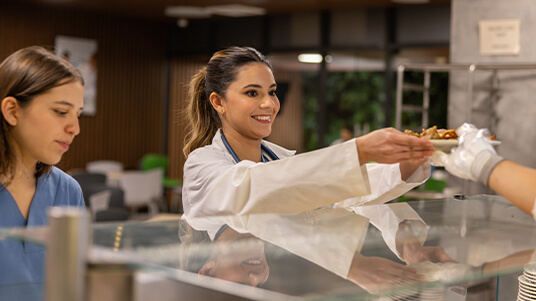 Healthcare workers at a cafeteria counter receiving food from a staff member behind the glass.