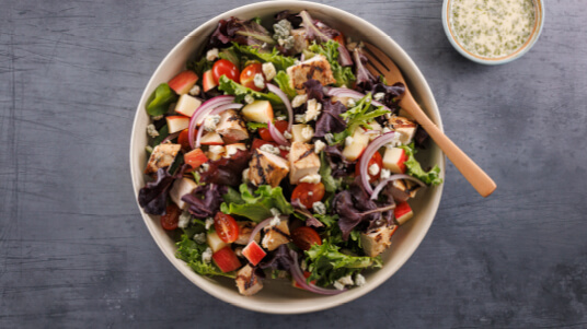 Bowl of mixed greens with grilled chicken, tomatoes, apples, and red onions beside a small cup of dressing.