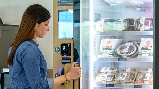 Person selecting fresh food from a refrigerated smart vending machine using a touchscreen payment panel.
