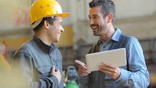 Two workers in an industrial setting discussing information on a tablet, one wearing a safety helmet.