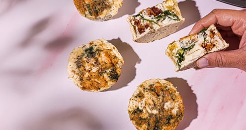 Assorted savory baked snacks arranged on a light pink surface, including herb topped pastries and sliced filled sandwiches, with a hand holding one piece in the corner of the image.Assorted savory baked snacks arranged on a light pink surface, including herb topped pastries and sliced filled sandwiches, with a hand holding one piece in the corner of the image.