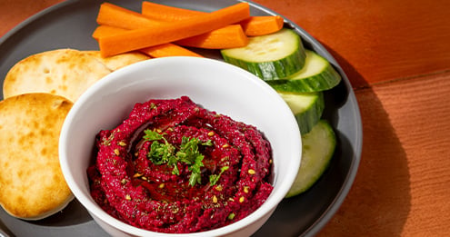 Bowl of bright beet dip garnished with herbs and olive oil, served with sliced cucumbers carrot sticks and pita bread on a dark plate placed on a wooden table.