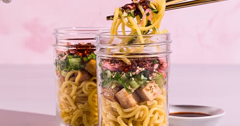 Two glass jars filled with noodle salad layered with vegetables tofu and seasonings, with chopsticks lifting noodles from one jar and a small dish of sauce nearby on a pink background.