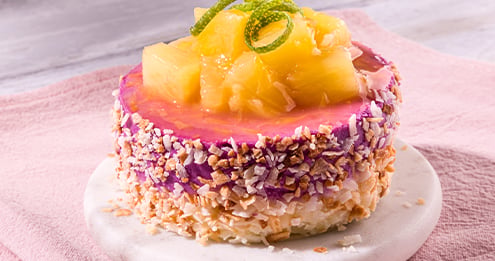 Round layered snack topped with pineapple pieces and citrus zest, coated in shredded coconut and chopped nuts, served on a small marble plate against a soft pink background.