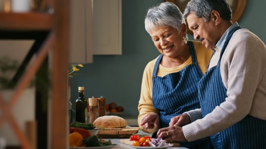 Two people wearing aprons preparing chopped vegetables together in a kitchen.