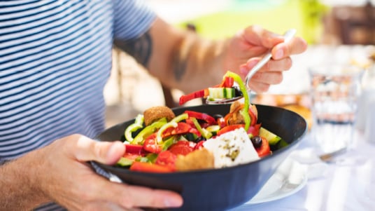 Person holding a bowl of fresh salad with tomatoes, cucumbers, peppers, and feta while eating with a spoon.