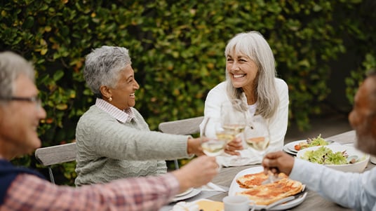People raising glasses and sharing a meal outdoors at a table surrounded by greenery.