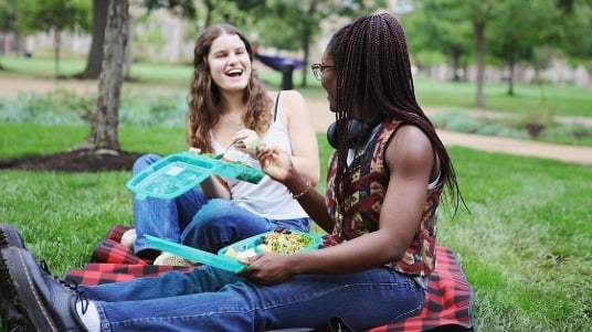 Two friends sit on a checkered blanket in a park, sharing a meal from reusable containers and smiling as they chat. 