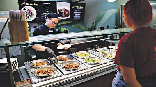 Dining hall staff places food into a bowl behind the counter while a student waits at a SUNY Oneonta dining hall station.