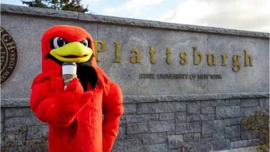 Burghy, the mascot of SUNY Plattsburgh, standing in front of the campus sign holding a cup of Saxbys coffee.