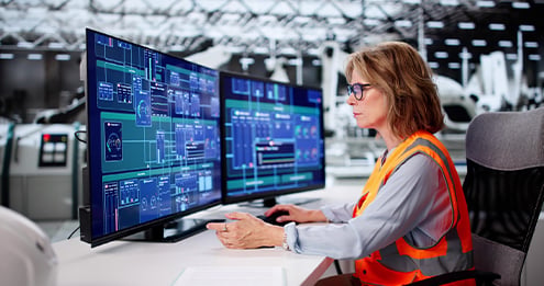 Facilities management professional monitoring operational dashboards across multiple screens in an industrial control room, overseeing building systems, performance data, and facility operations.