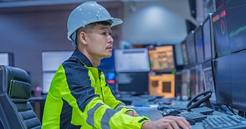 Industrial operations specialist wearing safety gear and a hard hat while monitoring data across multiple control room screens, highlighting facility management, process control, and operational performance.