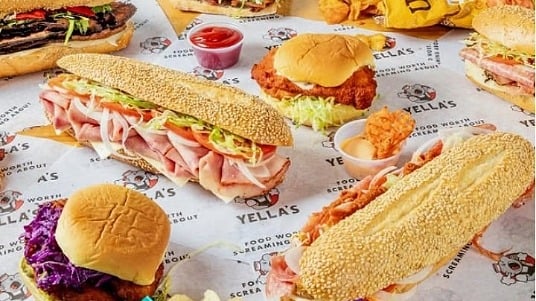 An assortment of Yella sandwiches, chips and waffle fries arranged on branded paper.
