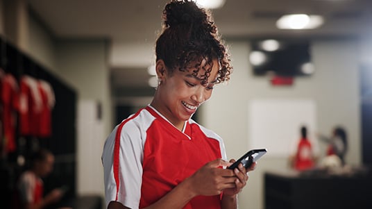 Person in a red athletic jersey using a smartphone while standing inside a sports locker room, with team uniforms, benches, and training equipment visible in the background.