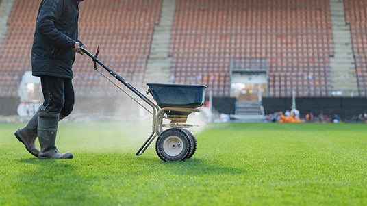 Grounds maintenance worker pushing a spreader across a sports field while applying lawn treatment, with stadium seating visible in the background.