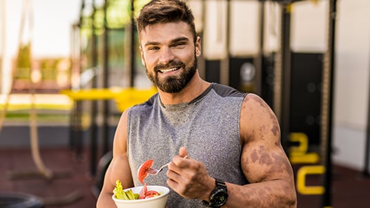 Athlete seated outdoors near a fitness training area holding a bowl of salad and a fork, with exercise equipment visible in the background during daylight.