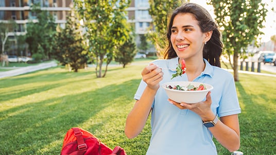 Person sitting on grass in a park, holding a bowl of salad and a fork, with buildings and trees behind.