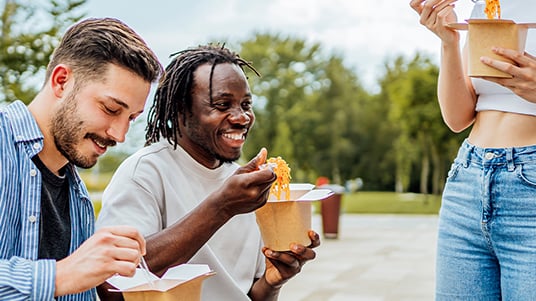 Three people outdoors sharing meals from takeaway containers at a table near a green open space, with trees and a paved gathering area visible in the background.