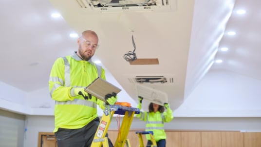 Facilities maintenance workers wearing high visibility safety clothing installing ceiling panels while standing on ladders in a bright indoor space with exposed wiring and lighting fixtures visible overhead.