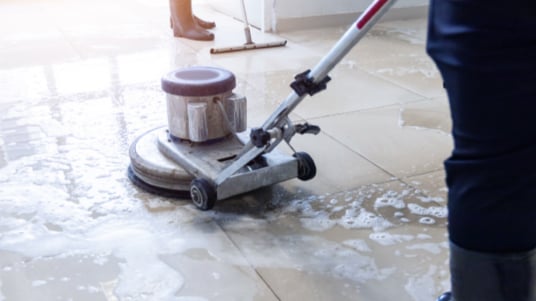 Floor cleaning machine scrubbing a tiled surface covered with water and cleaning solution, while another cleaning tool is visible in the background of a bright indoor facility.
