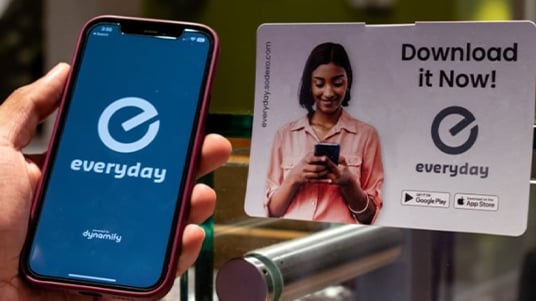 Close-up of a smartphone displaying the Everyday mobile app beside a promotional sign encouraging app downloads, featuring app store icons and a person using a mobile device.