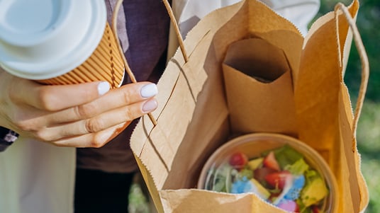 Close-up of a person holding a takeaway coffee cup and paper food bags, with a fresh salad in a compostable container visible inside, highlighting grab-and-go dining and meal pickup.