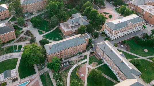 Copilot said: Aerial view of a college or university campus with multiple academic buildings, landscaped green spaces, walking paths, courtyards, and tree-lined grounds arranged across a large outdoor setting.