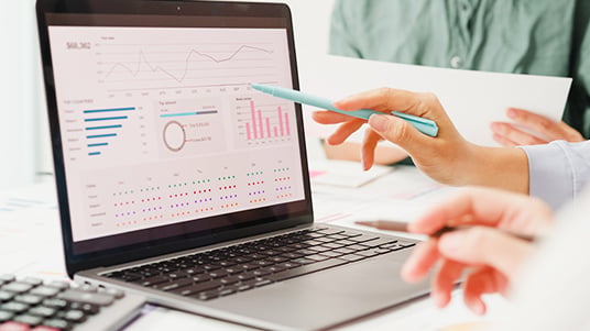 Close up of a laptop displaying charts graphs and analytics dashboards while people review printed documents and discuss data at a desk in a professional office setting.