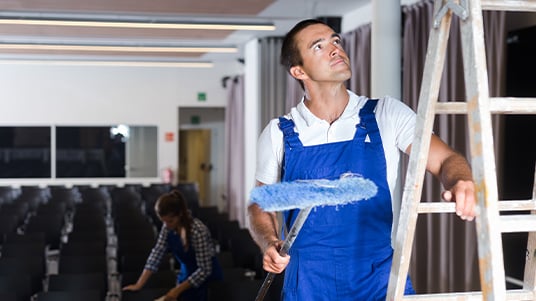 Facilities worker painting a ladder in a meeting room while another worker cleans nearby.