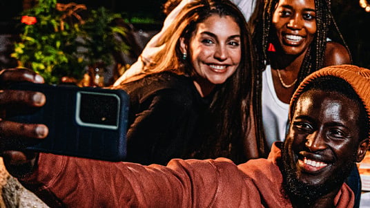 Group of people gathered outdoors at night taking a smartphone photo together, with string lights, greenery, and a social dining setting visible in the background.
