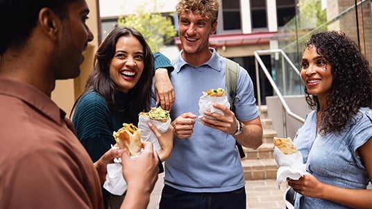 Group of people gathered outdoors on a campus walkway, holding wraps and sandwiches while talking together in a casual social setting with buildings and trees in the background.
