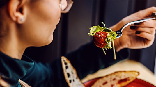 Close-up of a person holding a forkful of salad above a bowl filled with greens, vegetables, and toppings, with a wrap sandwich on the side in a casual dining setting.
