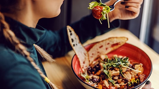 Close-up of a person holding a forkful of salad above a bowl filled with greens, vegetables, and toppings, with a wrap sandwich on the side in a casual dining setting.