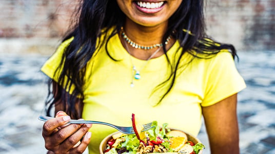 Close-up of a person holding a fork above a bowl of fresh salad with leafy greens, sliced vegetables, and colorful toppings, seated outdoors in a casual dining setting.